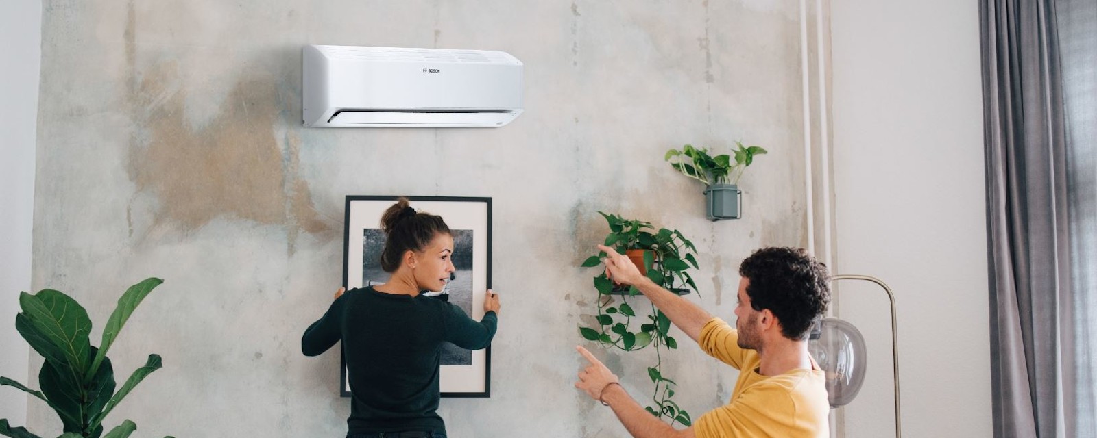 Bosch Split Klimagerät Climate Class 6000i hängend im Wohnzimmer, während eine Frau und ein Mann ein Bild darunter aufhängen.