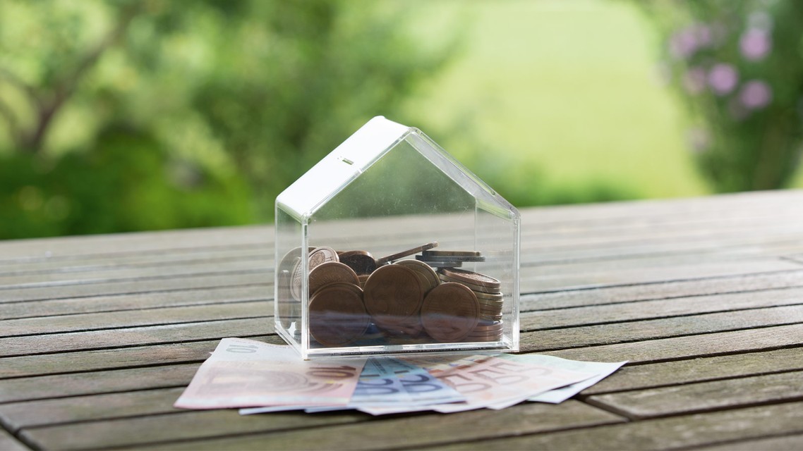 Spardose in Form eines Hauses steht auf einem Gartentisch und ist mit Geld gefüllt, unter Spardose sind Geldscheine geklemmt 
